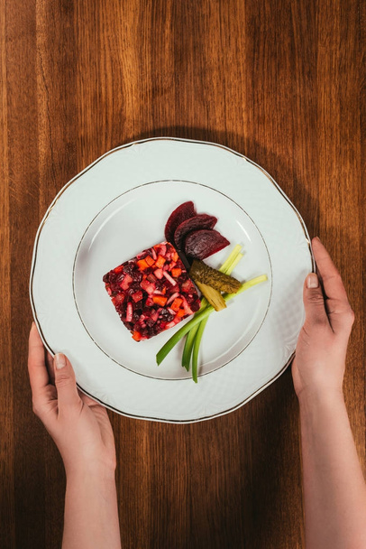 Top view of female hands holding plate with Vinegret salad served with green onion and pickled cucumber on wooden table - Valokuva, kuva