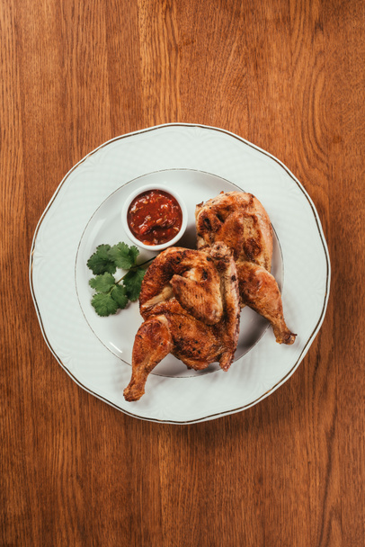 grilled chicken laying on plate with red sauce in saucer over wooden surface - Фото, зображення