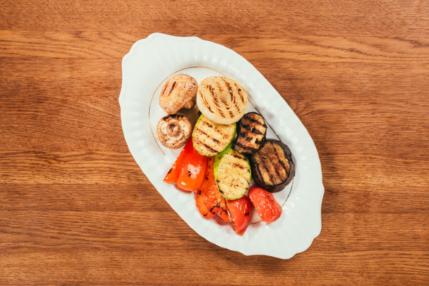 Top view of grilled vegetables and mushrooms on white plate on wooden table - Фото, изображение