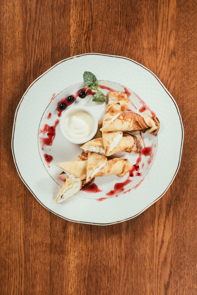 Bovenaanzicht van zoete crêpe pannenkoeken met jam en sour-cream op plaat op houten tafel - Foto, afbeelding