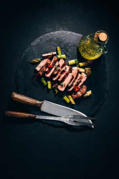vista superior de carne grelhada fatiada gourmet com espargos, óleo, garfo de carne e faca em placa de ardósia preta
  - Foto, Imagem