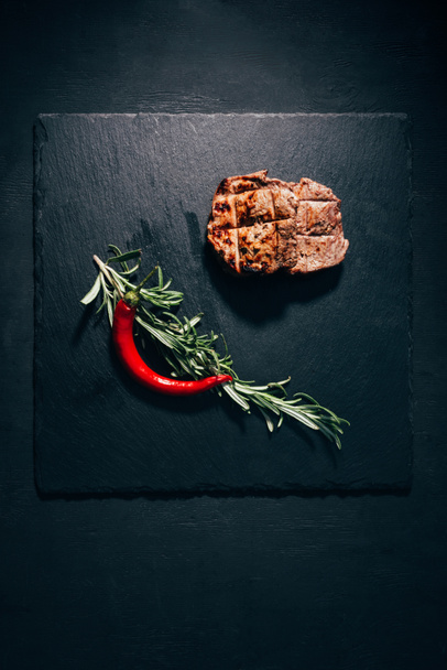 top view of gourmet grilled steak with rosemary and chili pepper on black slate board - Fotografie, Obrázek
