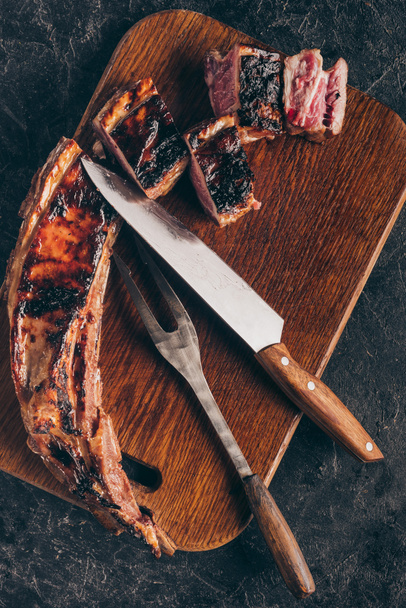 top view of delicious grilled meat with fork and knife on wooden board on black  - 写真・画像