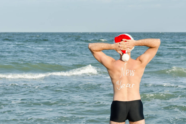 Der Mann mit dem Weihnachtsmannhut steht am Ufer des Meeres. Hände hinter dem Kopf - Foto, Bild