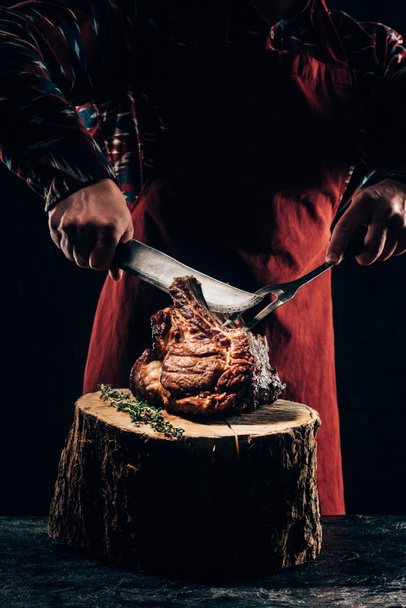 abgeschnittener Schuss von Koch in Schürze mit Gabel und Messer beim Schneiden köstlicher gegrillter Rippen auf Holzstumpf - Foto, Bild
