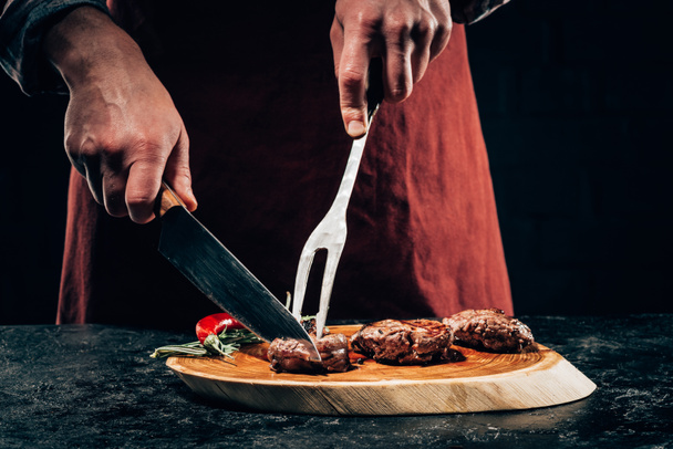 περικοπεί shot σεφ στην ποδιά με πιρούνι το κρέας και το μαχαίρι σε φέτες στη σχάρα γκουρμέ μπριζόλες με δεντρολίβανο και πιπέρι τσίλι σε ξύλινη σανίδα  - Φωτογραφία, εικόνα