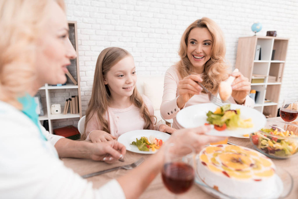 Mom, grandmother and little girl are dining together at home. They have vegetables and sweets on their table. - Фото, зображення