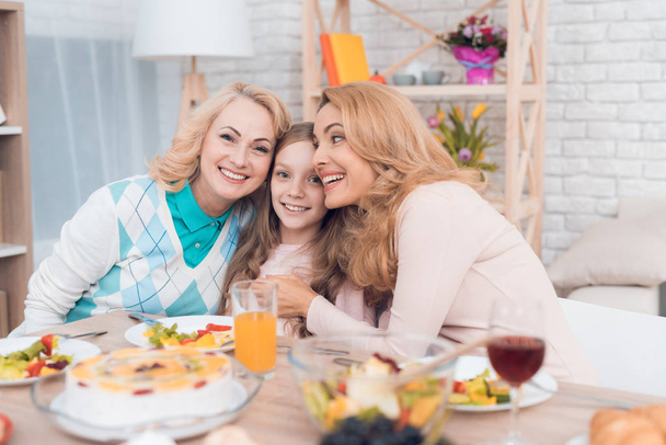 Two adult women and a girl are hugging, sitting at a table. This is the mother and grandmother of the girl. They have dinner at home. - Foto, Imagem