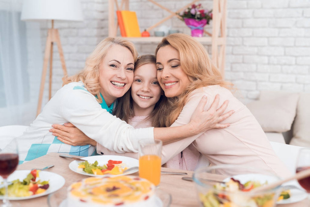 Two adult women and a girl are hugging, sitting at a table. This is the mother and grandmother of the girl. They have dinner at home. - Foto, afbeelding