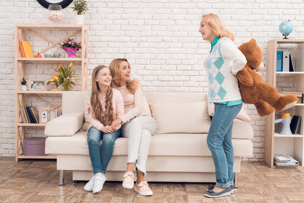 Mom and grandmother give a little girl a big toy bear. They make her a surprise. The family has dinner at home - they have a holiday. - 写真・画像