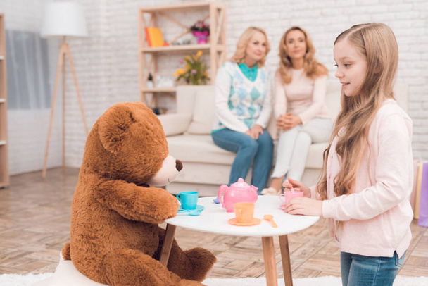 Little girl is playing with a toy bear. There are toy cups on the toy table. Behind sit her mother and grandmother. - Foto, afbeelding