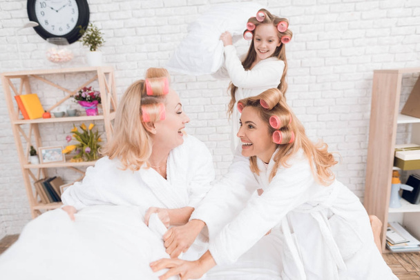 Two adult women and a girl play a fight with pillows on the bed. They have curlers on their heads. They are in a good mood. - Valokuva, kuva