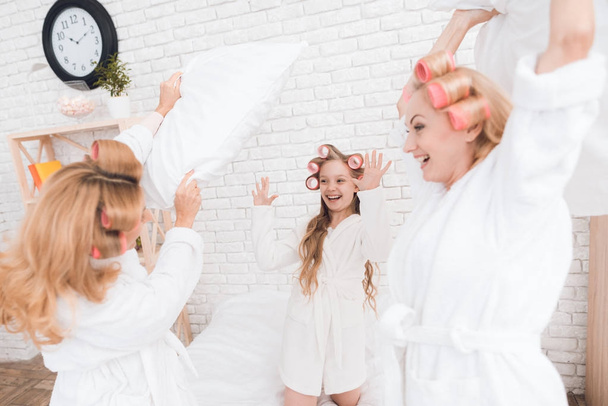 Two adult women and a girl play a fight with pillows on the bed. They have curlers on their heads. They are in a good mood. - Foto, Bild