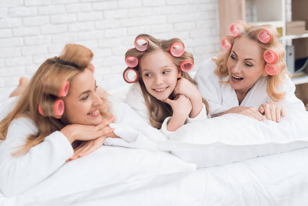 Two adult women and a girl are lying on the bed. They have curlers on their heads. They are in a good mood. - Fotoğraf, Görsel