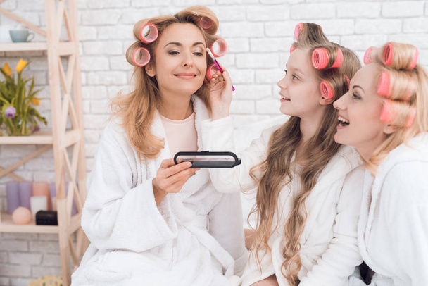 Mom, grandmother and girl in curlers make each other make-up. They sit on a bed in a bathrobe. They are in a good mood. - Photo, image