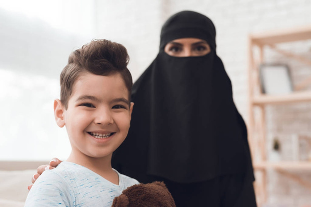 A little boy from an arab family is holding a toy bear in his hands. This is a traditional muslim family at home. The boys mother wears a parandja. - Photo, image