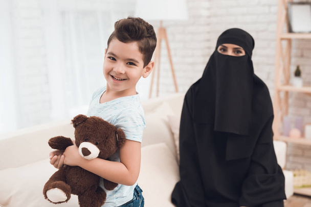 Un niño de una familia árabe sostiene un oso de juguete en sus manos. Esta es una familia musulmana tradicional en casa. La madre de los chicos lleva una parandja
. - Foto, Imagen