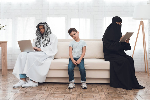 A boy from a traditional Arab family is angry with parents who do not notice him. A woman and a man are looking at the tablet and laptop. They sit together on the couch. - Foto, Imagen
