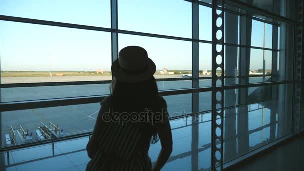 Молода жінка в капелюсі з рюкзаком приходить у вікно терміналу аеропорту і дивиться на злітно-посадкову смугу. Дівчина чекає на свій літак. Туризм і концепція подорожей. Вид ззаду Повільний рух
 - Кадри, відео