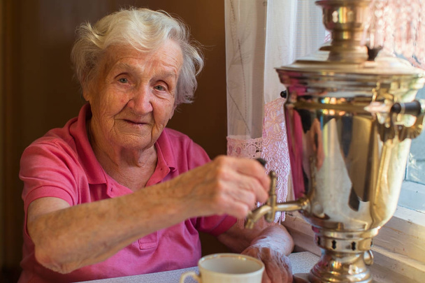 Eine ältere Frau trinkt Tee am Tisch. klassischer russischer Samowar. - Foto, Bild
