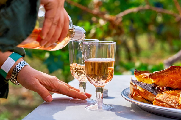 Female hands pour refreshing pink wine in a glass with cakes on the table, against the background of the vineyard. the concept of winemaking. - Photo, Image