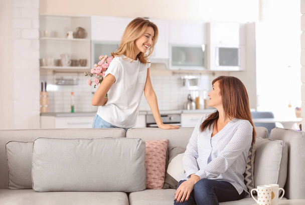 Attractive young woman hiding bouquet for mother behind her back at home - Foto, Bild