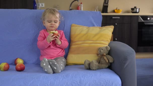 Lovely baby girl sitting on the sofa and eating big apple fruit. - Imágenes, Vídeo
