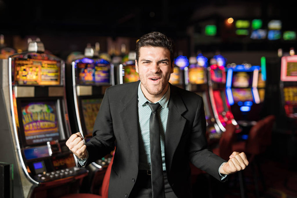 Vonzó fiatal férfi keres izgatott, ütő a jackpot egy slot machine egy kaszinóban öltöny - Fotó, kép