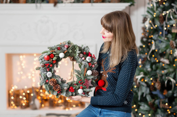 Young woman holding a Christmas wreath with fir branches for the holiday. The new year celebration. on the background of fireplace and Christmas tree, lights, garlands - Фото, зображення