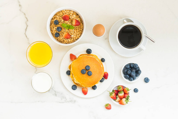 gesundes Frühstücksset mit Pfannkuchen und Müsli mit Blaubeere und Erdbeere und schwarzem Kaffee, Milch und Orangensaft auf weißem Steintischhintergrund - Foto, Bild