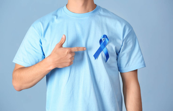 Young man pointing at blue ribbon on his t-shirt against color background, closeup. Prostate cancer awareness concept - Photo, image