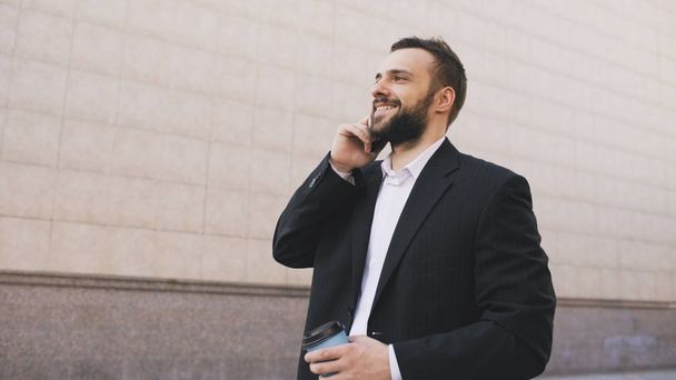 Young bearded business man talking on cell phone making deals with cup of coffee near modern office buildings - Photo, Image