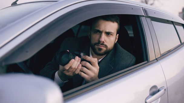 Young private detective man sitting inside car and photographing with dslr camera - Фото, изображение