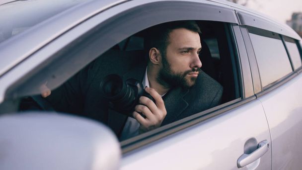 Young private detective man sitting inside car and photographing with dslr camera - Zdjęcie, obraz