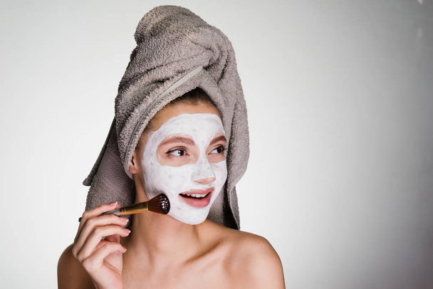 attractive young girl with a towel on her head puts a white mask on her face with a brush - Photo, Image