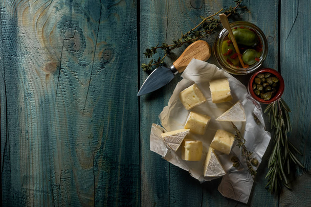 acostado plano con queso brie, cuchillo, hierbas y frascos con aceitunas y capperi en la mesa rústica azul
 - Foto, Imagen