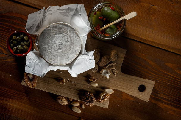 flat lay with brie cheese, knife, herbs and jars with olives and capperi on rustic wooden table - Фото, изображение