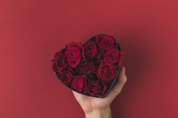 tiro recortado de hombre sosteniendo rosas en caja en forma de corazón en la mesa roja, San Valentín concepto de día
 - Foto, Imagen