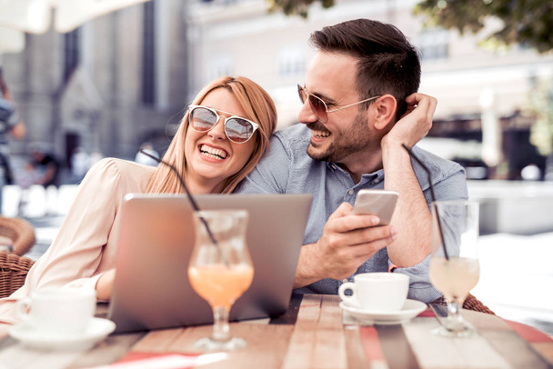 Szczęśliwa para siedzi w ulicznej kawiarni razem i za pomocą laptopa i telefonu. - Zdjęcie, obraz
