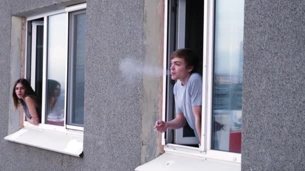 Jonge man gesprekken met een buurman door het raam. Jonge man rookt een sigaret en praat met een buurman van zijn raam - Video