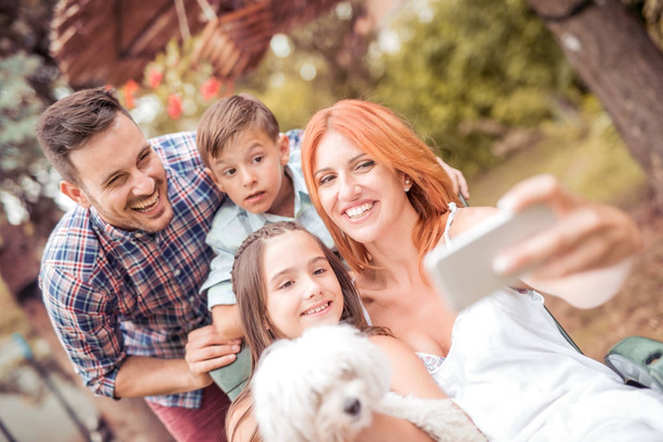 Famille heureuse avec chien prenant selfie avec téléphone intelligent dans le parc.Famille, animal de compagnie, technologie et concept de personnes
. - Photo, image