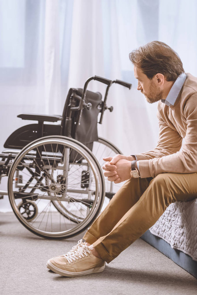 worried man with disability sitting on bed and looking at wheelchair - Foto, Imagen