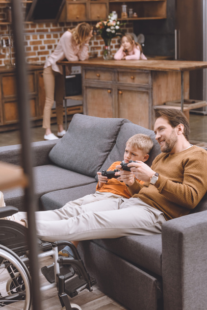 selective focus of happy disabled father with legs on wheelchair and cute little son playing with joysticks together at home  - Fotoğraf, Görsel