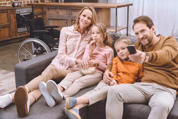 ευτυχισμένη οικογένεια κάθεται στον καναπέ και βλέποντας τηλεόραση μαζί και αναπηρική καρέκλα στέκεται πίσω από - Φωτογραφία, εικόνα
