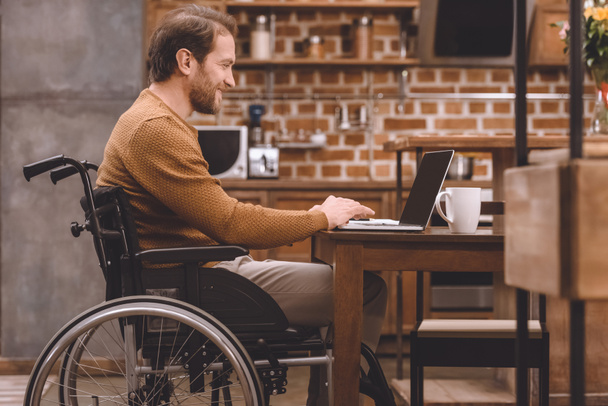 вид сбоку улыбающегося инвалида в инвалидной коляске, пользующегося ноутбуком дома
 - Фото, изображение