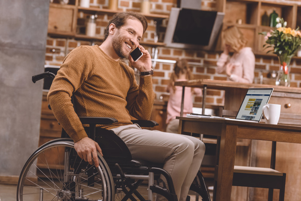 smiling disabled man in wheelchair talking on smartphone and using laptop at home - Foto, immagini
