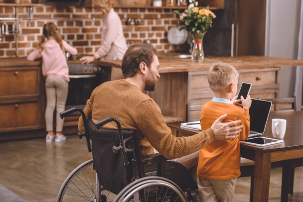 назад вид отца-инвалида в инвалидной коляске и маленького сына с помощью смартфона и ноутбука вместе на дому
 - Фото, изображение