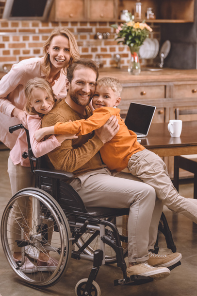mutlu aile babası sarılma ve evde kameraya gülümseyen tekerlekli sandalyede olan iki çocuk - Fotoğraf, Görsel