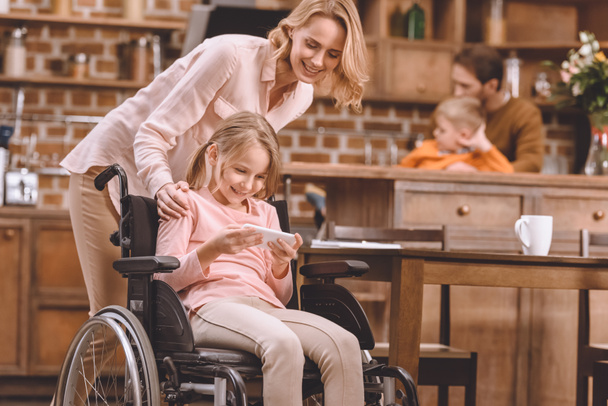 ευτυχισμένη μητέρα βλέπει χαμογελώντας ανάπηρη κόρη κάθεται σε αναπηρικό καροτσάκι και χρησιμοποιώντας το smartphone στο σπίτι  - Φωτογραφία, εικόνα