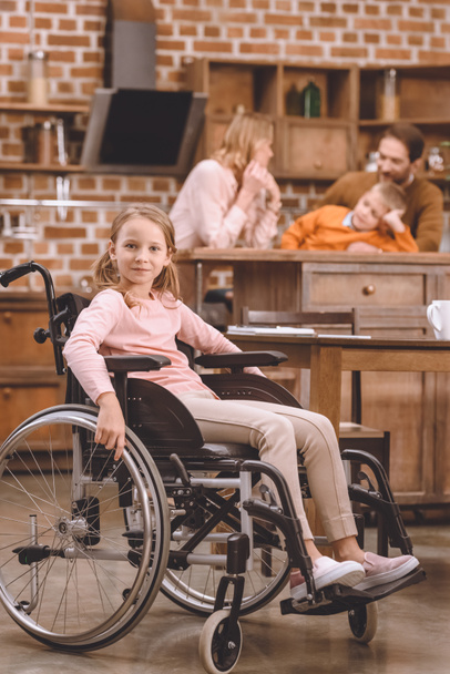 linda niña en silla de ruedas mirando a la cámara mientras otros miembros de la familia de pie detrás de casa
 - Foto, Imagen